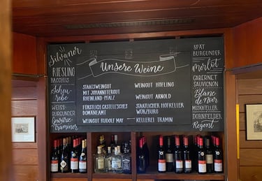 Handwritten chalk wine menu listing Silvaner, Riesling, and Merlot above a shelf of bottled German wines.