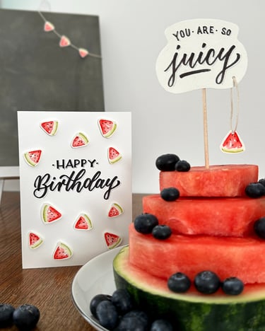 Handmade watermelon birthday card next to a tiered fresh watermelon cake decorated with blueberries.