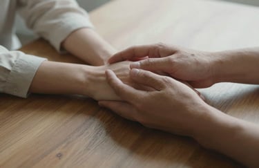 A close-up shot of two people's hands resting on a wooden table, one offering support to the other, captured in soft natural light with soft off-white tones.