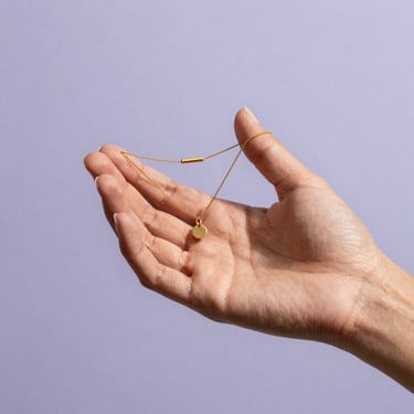 A woman's hand holding a delicate piece of minimalist gold jewelry against a light lilac backdrop, South American vibe.