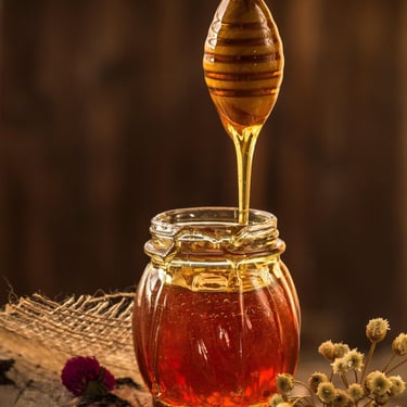 Golden organic honey dripping from a wooden dipper into a glass jar on a rustic table.
