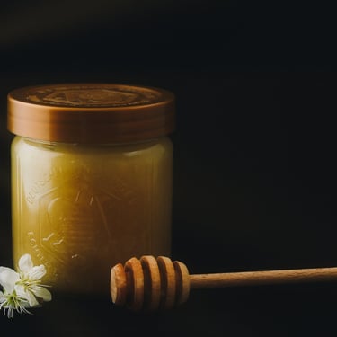 A glass jar of organic raw honey next to a wooden honey dipper and white flowers.