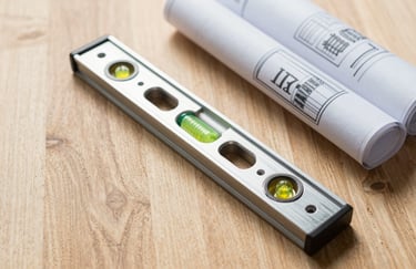 A flat lay photograph of professional construction tools, including a metallic level and rolled blueprints on a light oak wooden surface in a North American office.