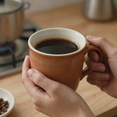 A close-up shot of a cozy hand-thrown terracotta ceramic mug filled with rich coffee, held with both hands in a warm, welcoming home kitchen.