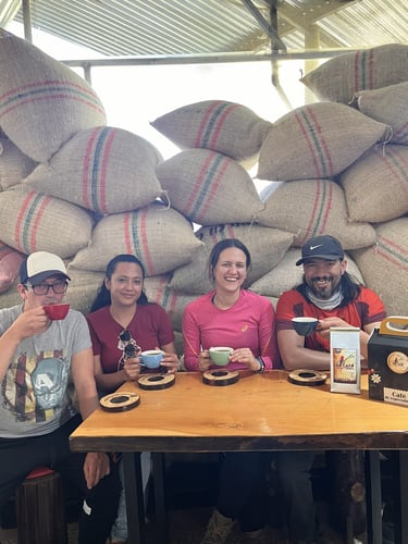 Friends tasting local coffee at a wooden table in front of burlap sacks of fresh coffee beans.