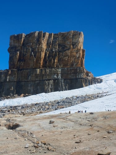 Hikers trekking past a massive rock formation and a snowy glacier under a clear blue sky.