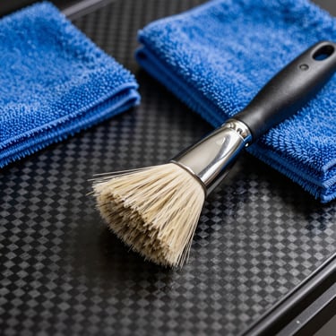 A close-up of professional automotive detailing brushes and electric blue microfiber cloths resting on a black carbon fiber panel, International / Global.