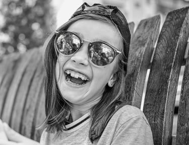 Uśmiechnięta dziewczynka w okularach przeciwsłonecznych, z czapką z daszkiem założoną tyłem.