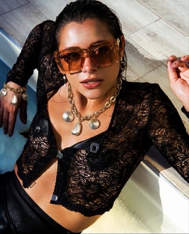 Woman in a black lace top and tinted sunglasses wears chunky gold statement jewelry by a pool.