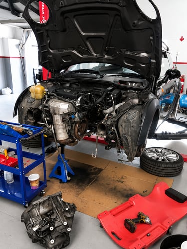 A car with an open hood undergoing engine repair at an auto service shop with tools and a jack stand.
