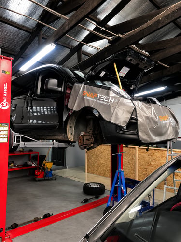 Black car on a red Mac Afric hydraulic two-post lift in an auto repair shop with Snaptech equipment.