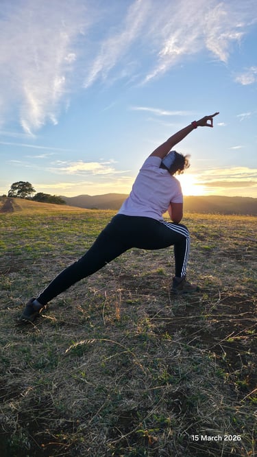 A woman practices outdoor yoga in a side lunge pose at sunset on a grassy hilltop.