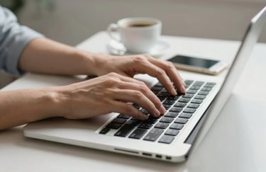 A close-up of human hands typing on a sleek silver laptop in a bright environment. On the desk is a white smartphone and a cup of coffee. Professional Central European style.