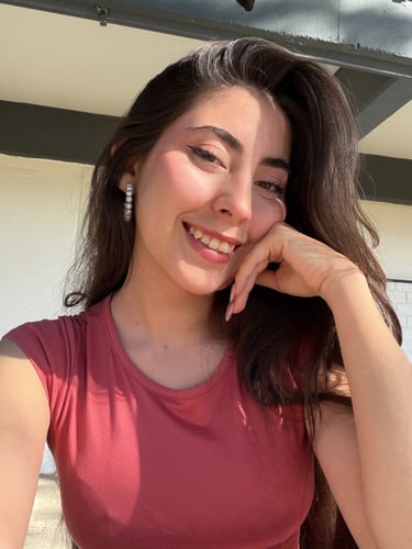 Mujer sonriente con cabello largo y oscuro y aretes de perlas, vistiendo una camiseta rosa