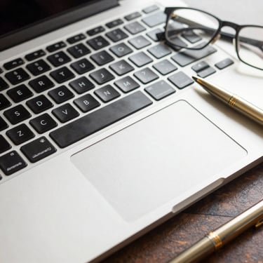 Macro photography of a sleek laptop keyboard and trackpad in a sunlit North American / US office, with a pair of professional glasses and a gold pen nearby.