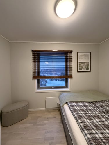 Cozy minimalist bedroom with a plaid duvet, wooden window blinds, and a gray ottoman on light wood floors.