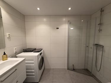 Modern white tiled bathroom featuring a walk-in glass shower, vanity sink, and front-load washing machine.