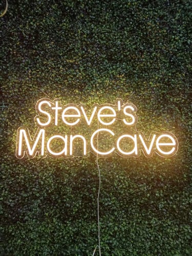 LED Neon Schild „Steve's ManCave“ in warmweiß auf grüner Pflanzenwand