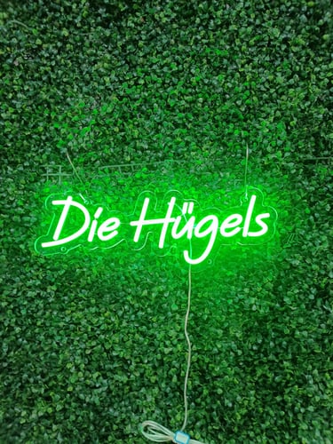 grünes LED Neon Schild mit Schriftzug „Die Hügels“ auf grüner Pflanzenwand