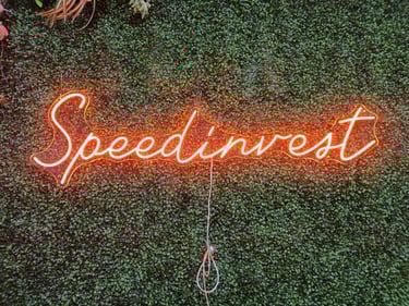 oranges LED Neon Schild mit Schriftzug „Speedinvest“ auf grüner Pflanzenwand