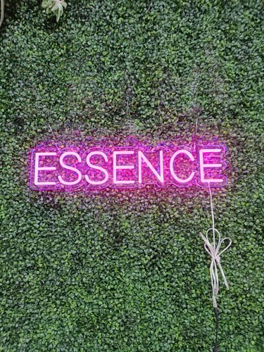 pinkes LED Neon Schild mit Schriftzug „ESSENCE“ auf grüner Pflanzenwand