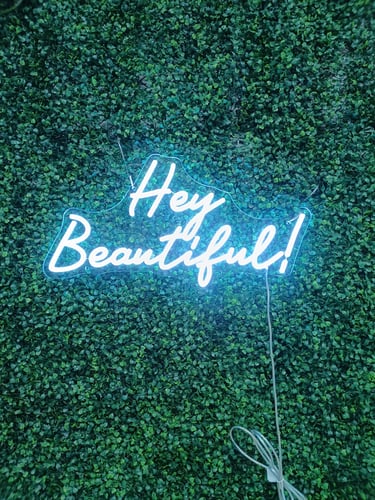 kaltweißes LED Neon Schild mit Schriftzug „Hey Beautiful!“ auf grüner Pflanzenwand
