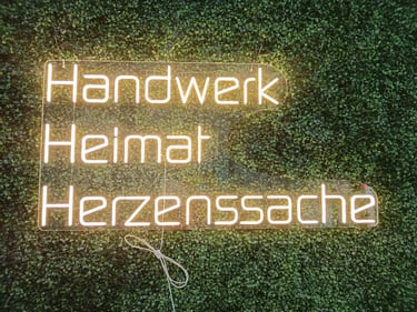 warmweißes LED Neon Schild mit Schriftzug „Handwerk Heimat Herzenssache“ auf grüner Pflanzenwand in einer Bäckerei