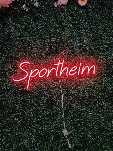 rotes LED Neon Schild mit Schriftzug „Sportheim“ auf grüner Pflanzenwand