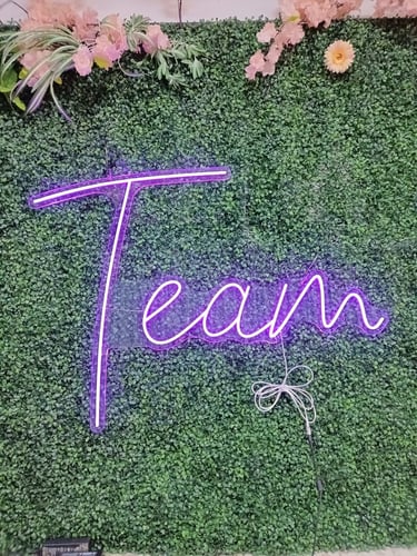 lila LED Neon Schriftzug „Team“ auf grüner Pflanzenwand mit dekorativen Blumen