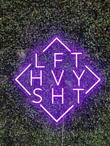 lila LED Neon Logo mit geometrischem Rahmen und Buchstaben LFTHVYSHT auf grüner Pflanzenwand