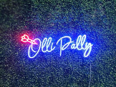 blaues LED Neon Schild Olly Pally Schriftzug mit Pfeil als personalisierte Neon Dekoration auf Pflanzenwand