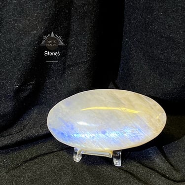 Polished moonstone palm stone with blue flash on a display stand against a black background.