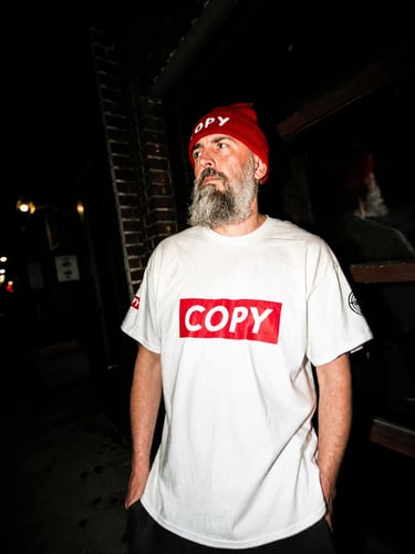  Dacarter in a red "COPY" beanie and white "COPY" t-shirt stands outside at night.