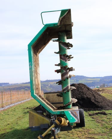 A Mechanical Compost Turners attachment machine stands near a compost pile in an agricultural field.