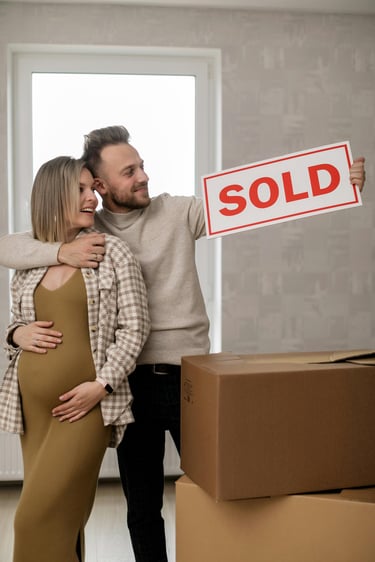 Happy pregnant couple holding a red sold sign while standing next to moving boxes in their new home.