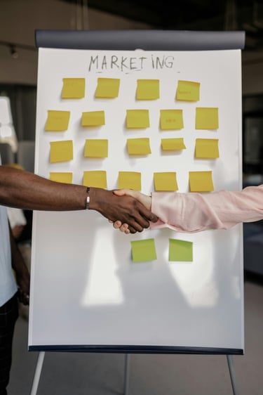 A handshake between business colleagues in front of a marketing strategy whiteboard with sticky notes.