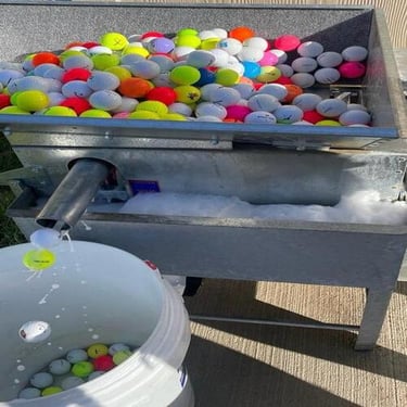 Automatic golf ball washer cleaning multi-colored golf balls into a white collection bucket.