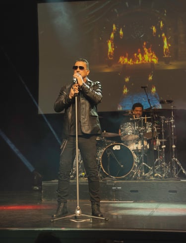 Male rock singer in a black leather jacket performing live on stage with a drummer and fire visuals.
