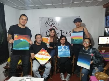 Personas sonrientes mostrando pinturas hechas durante un taller a mano en sala con luz cálida y pendón en la pared