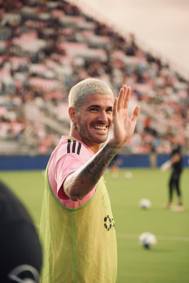 Rodrigo De Paul smiling and waving at fans during a soccer training session in a stadium.