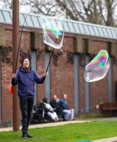 A man creates giant iridescent bubbles outdoors using a wand kit in a public park.
