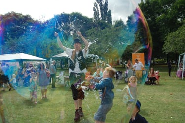 A street performer blowing giant soap bubbles for children at an outdoor summer festival in a park.