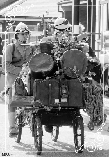 Vintage black and white photo of men in steampunk costumes riding a bicycle loaded with antique luggage.