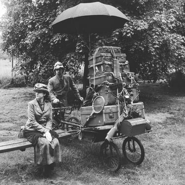 Vintage travelers in steampunk attire with a bicycle cart overloaded with antique luggage and umbrellas.