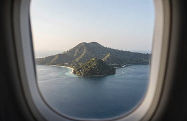 A travel-themed shot of a scenic Southeast Asian / Indonesian island landscape seen through a modern airplane window, soft morning light, Muted Steel Blue water.
