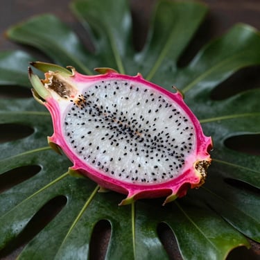 Halved vibrant pink pitahaya showing its white flesh and black seeds, resting on a dark green tropical leaf.