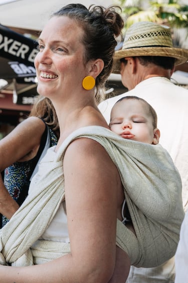 Smiling woman wearing a sleeping infant in a woven linen baby wrap carrier at an outdoor market.