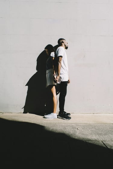 A young couple standing back-to-back holding hands against a white brick wall in sunlight.