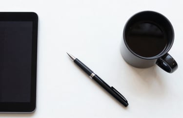 A top-down view of a designer's desk with a black coffee mug, a sleek tablet, and a professional digital pen on a white surface. The composition is clean, balanced, and symbolizes innovative digital strategy.