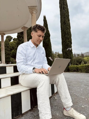 A man in a white shirt and linen pants working on a silver laptop outdoors in a park.